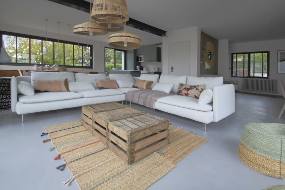 Salon moderne avec canapé blanc, table en bois et tapis tissé. Grandes fenêtres ouvertes.